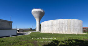 Tall concrete water tower with a yellow railing beside a curved cylindrical silo against a bright blue sky.