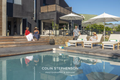 Two women sit on wooden steps by a dark modern house while a man stands near beige pool loungers under white umbrellas, on a sunny day.