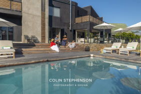Group of people relaxing and chatting on a wooden pool deck in front of a modern dark building with patio seating and umbrellas nearby.