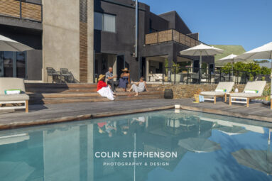 Group of people relaxing and chatting on a wooden pool deck in front of a modern dark building with patio seating and umbrellas nearby.