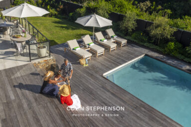 Aerial view of a poolside wooden deck with four beige lounge chairs under white umbrellas and a group of people chatting nearby with towels and hats on the deck.