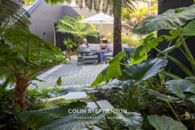 Two people sit on outdoor lounge chairs under a white umbrella in a lush courtyard garden with tropical plants.