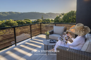 Terrace seating with wicker sofas and a reader in a wide-brim hat perusing a magazine, overlooking green hills at sunrise.