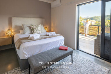 Bedroom with a large bed, decorative pillows, and night lamps; glass doors open to a balcony where a person in a striped dress stands overlooking hills.