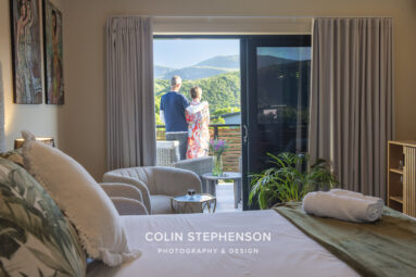 Couple standing on a balcony looking at green hills and mountains from a living room doorway, with curtains open and indoor furniture in view.