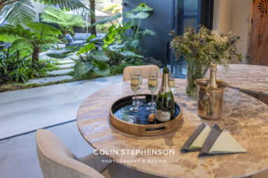 Indoor dining table with champagne on ice and four fluted glasses; open doors reveal a lush tropical garden with ferns and large green leaves in the background, plus a vase of white flowers on the table edge.