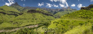 Panoramic view of green rolling hills and rocky mountains under a blue sky with white clouds, a sunlit landscape stretching to the horizon in the distance S