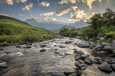 Rocks and a fast-flowing river in a valley with green hills and distant mountains under a blue, partly cloudy sky.
