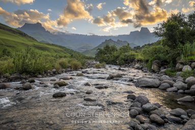 Rocky riverbed in the foreground winds through a green valley toward distant mountains under a golden, partly cloudy sunset sky.