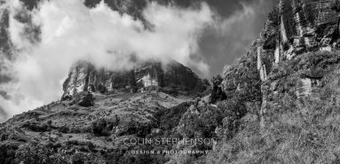 Black-and-white mountain landscape with steep rocky cliffs and cloudy sky, foreground vegetation visible, watermark at the bottom reads Colin Stephenson Design & Photography (text not essential to scene). Alternative text emphasizes the dramatic mountainous scene.