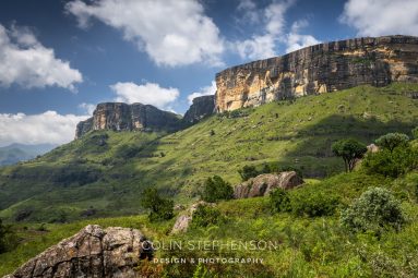Rocky cliff face rising above green hills under a blue sky with scattered white clouds.