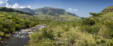 Panoramic view of a river winding through a green valley with grassy hills and distant rugged cliffs under a blue sky.