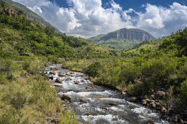 Wide river flows over rocks through a green valley with forested hills and distant mountains under a blue sky with clouds.