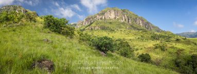 Panoramic view of green hills and rocky mountains under a blue sky with a few clouds.