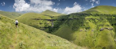 Hiker with backpack walking along a grassy hillside toward a valley with a small waterfall and green rolling hills under a blue sky.