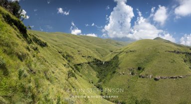 Wide panorama of green rolling hills under a bright blue sky with fluffy white clouds; rocky hillside on the left and grassy foreground