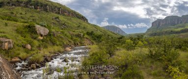Panoramic landscape of a grassy valley with a rocky hillside on the left, a rushing stream with rocks in the foreground, and distant cliffs under a cloudy sky.