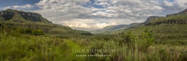 Panoramic view of a grassy valley flanked by green hills and distant cliffs under a cloud-filled sky.