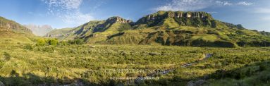 Panoramic view of a green valley with rolling hills and rugged cliffs under a clear blue sky, a small river winding through the foreground.